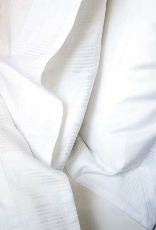 Photo by Kay Bowling of a Soleil Border on a pillowcase, Euro Sham, top sheet, and duvet cover.  Soleil border is an abstracted ray of the sun white on white jacquard weave.  The body of the linens is Soleil 300 thread count sateen.  Part of Kearsley Daily Essentials.